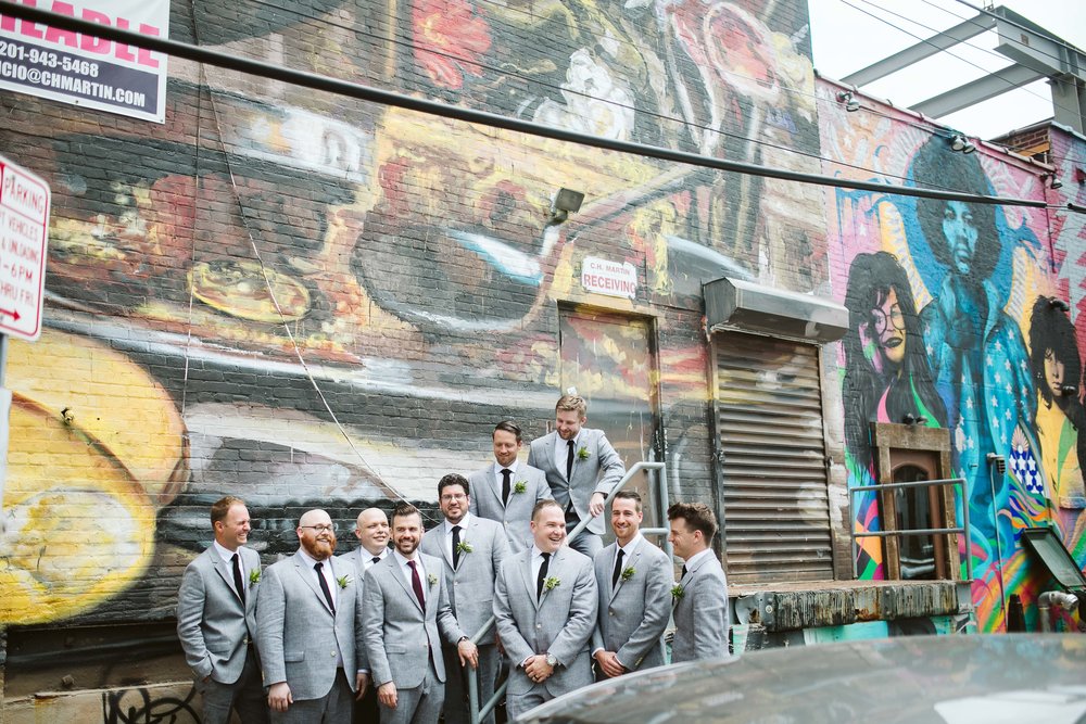  The groom and groomsman walking to the wedding in their gray suits by a graffiti wall at this Battello Wedding in Jersey City, NJ 