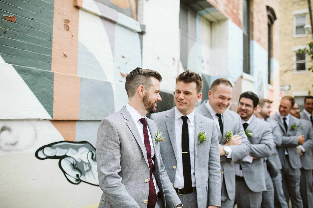 The groom and groomsman walking to the wedding in their gray suits by a graffiti wall at this Battello Wedding in Jersey City, NJ 